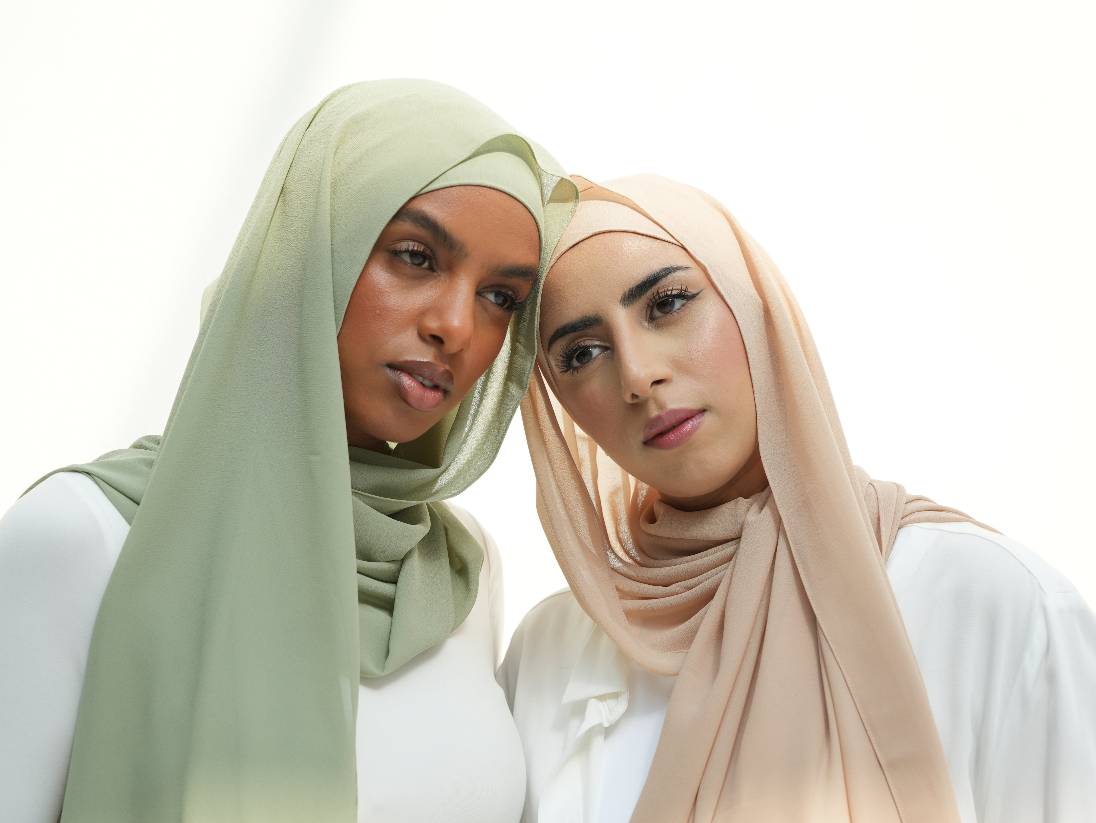 Two women wearing hijabs standing close together on a white background