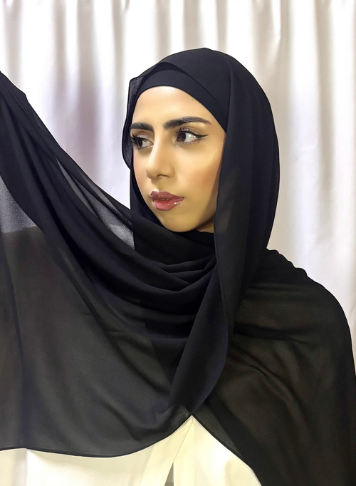 A woman wearing a black chiffon scarf with a non-slip undercap, in a room with curtains.